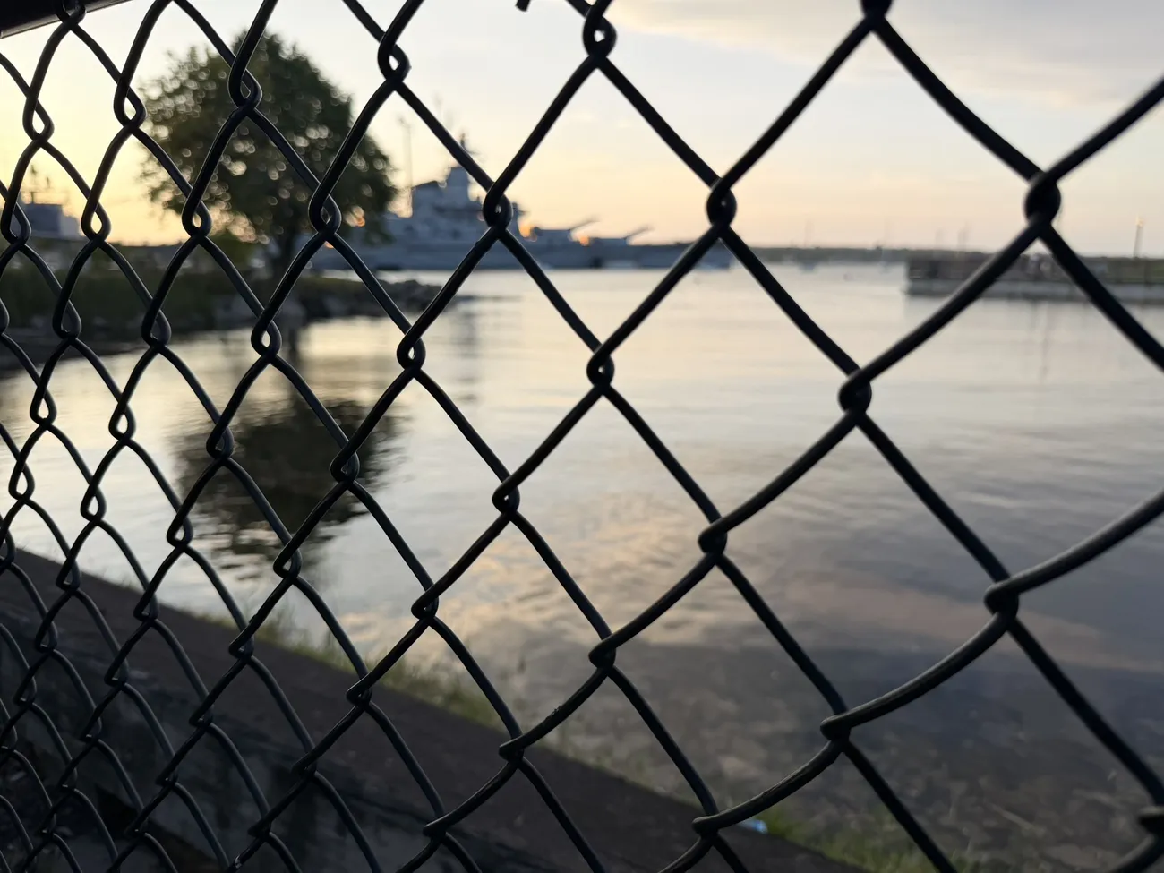 The battleship at Battleship Cove, seen through a chain-link fence at dawn.
