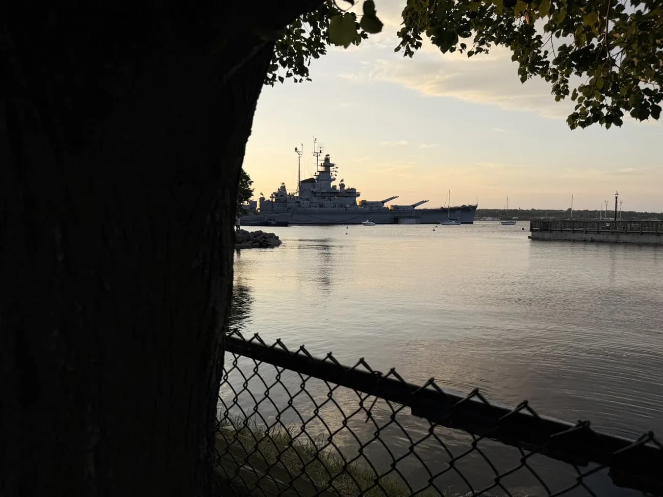 The battleship at Battleship Cove, seen through a chain-link fence at dawn.