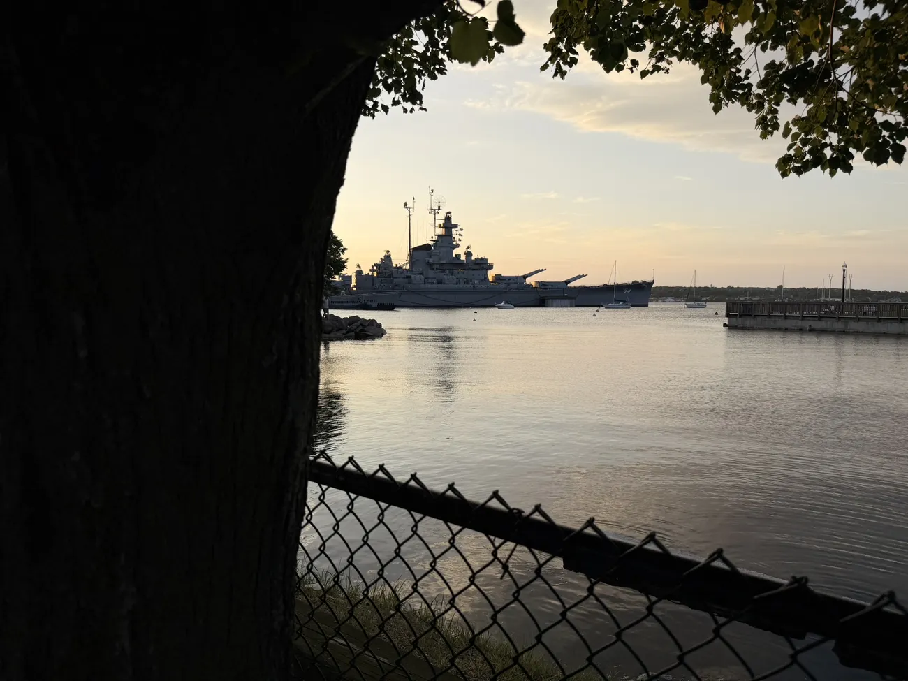 The battleship at Battleship Cove, seen through a chain-link fence at dawn.